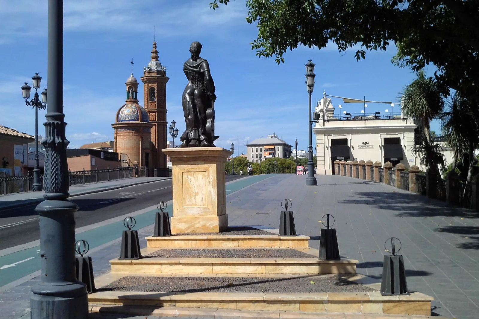 A bronze statue on a stone plinth in a square in Triana, with the bell tower of a small chapel and an iron bridge behind it