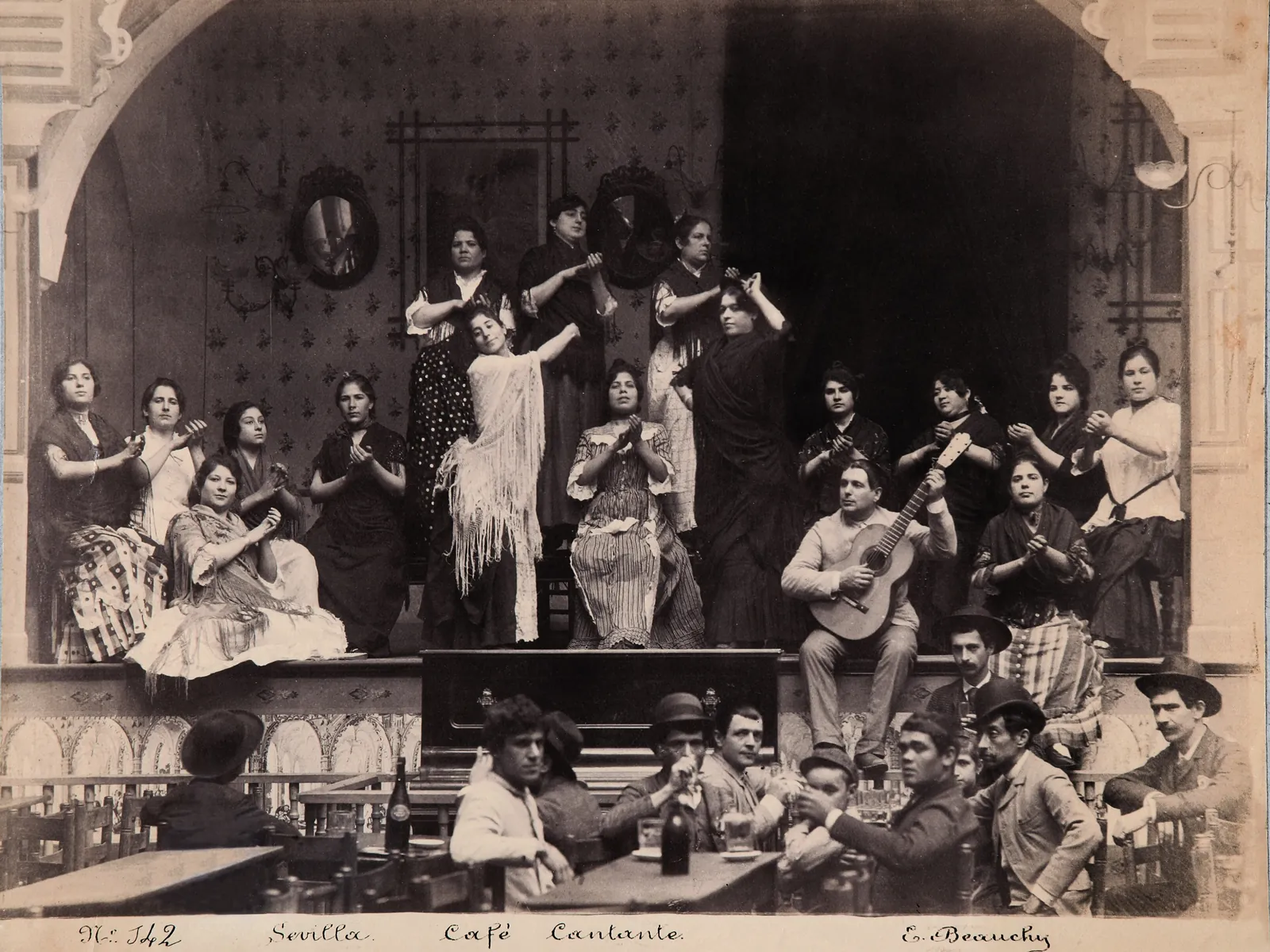 An old black-and-white photograph of a café cantante in Seville, with a group of women in shawls and men with guitars posed on a small stage