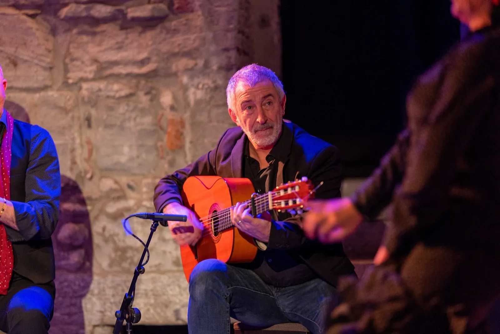 A flamenco guitarist seated on a stage, playing an orange-topped guitar into a microphone, with another performer in the foreground