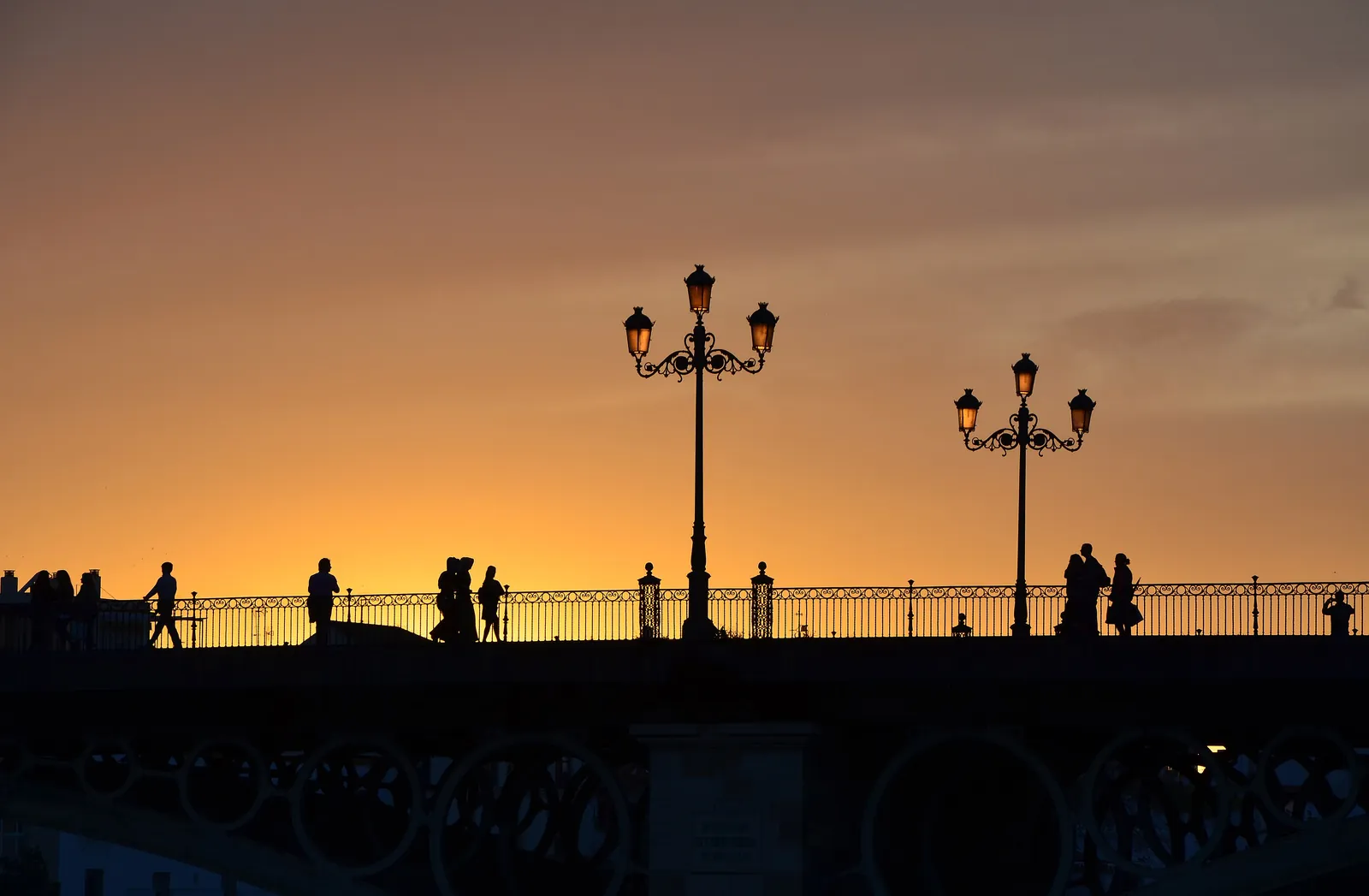 The Triana Bridge at dusk, two street lamps and walkers silhouetted against an orange sky