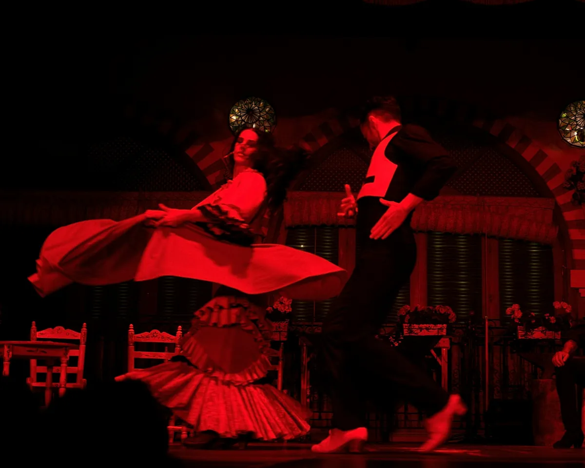 A flamenco dancer in a red dress and a male dancer in black on a tablao stage lit in red, two performers seated at a table on the right
